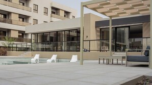 Un área de piscina moderna al aire libre con sillones de descanso, una zona de estar techada y un edificio con balcones y ventanas amplias.