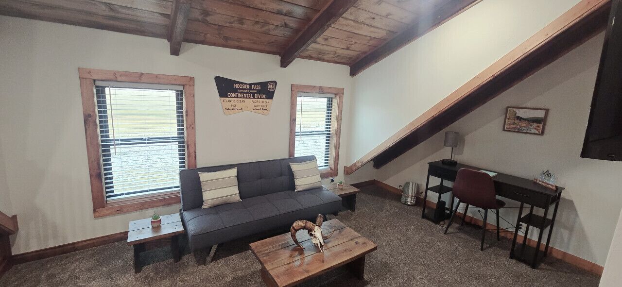 Habitación tipo loft con sofá, una mesita, escritorio y dos ventanas con persianas.