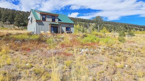 Una cabana de fusta de dos pisos amb teulada verda, situada en un camp verd amb un turó forestal al fons.
