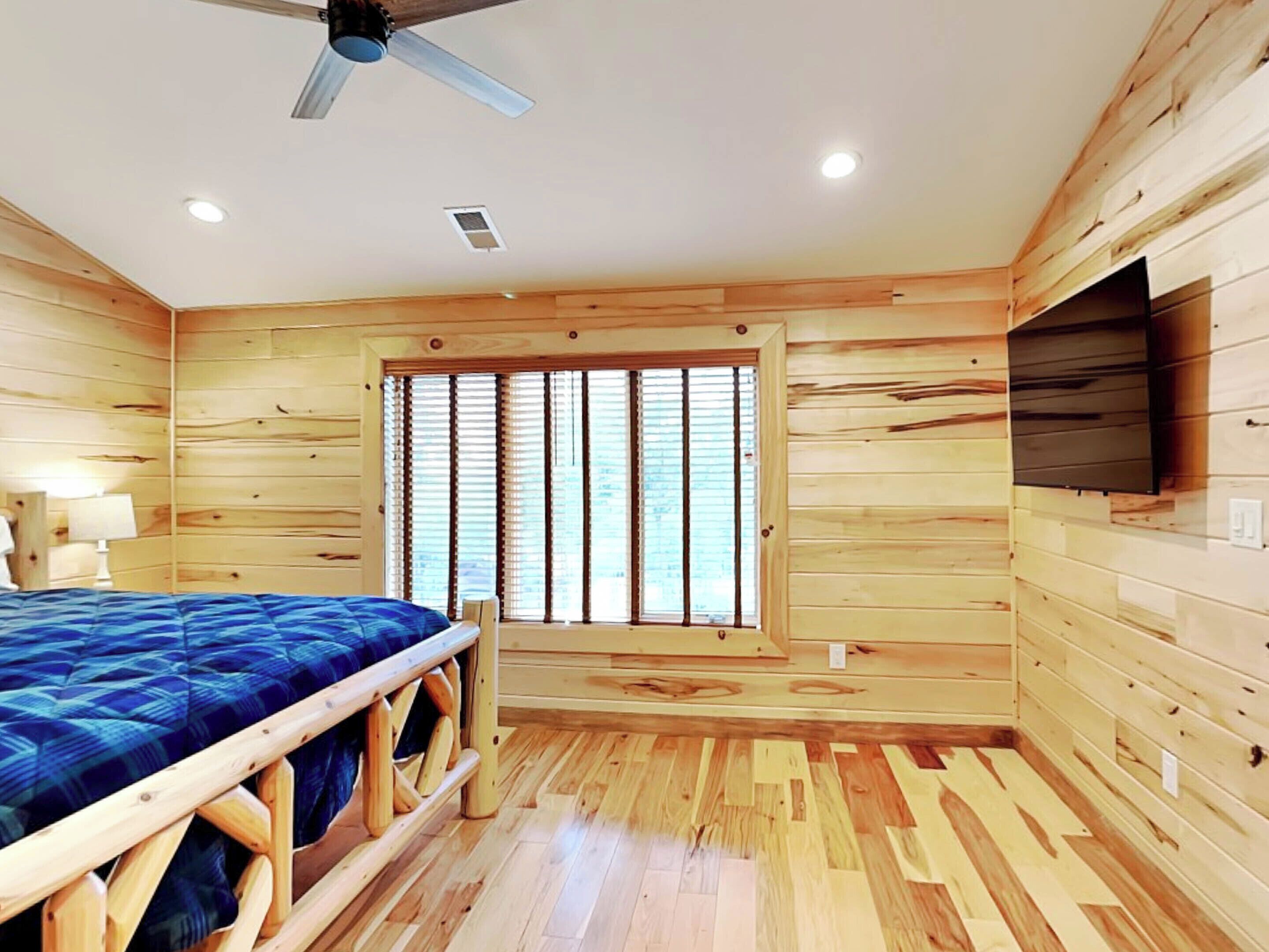 Ein Zimmer mit Holzwänden, einem Bett, einem Deckenventilator, einem Fenster mit Jalousien und einem angebrachten Fernseher.