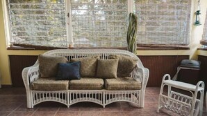 A wicker sofa with cushions, a small white metal bench, and a window with blinds. - Villa del Rio Room #1

 (Luquillo)
