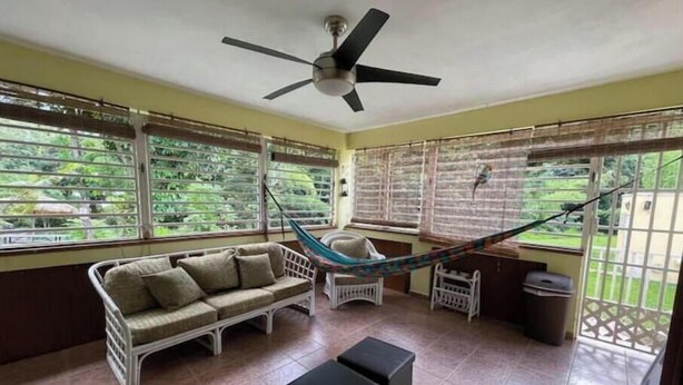 A room with a hammock, a sofa, a small table, and large windows with blinds. - Villa del Rio Room #1

 (Luquillo)