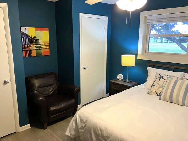 A bedroom with a bed, a chair, a nightstand, a lamp, a painting, and a door. - Surfside Beach Bungalow- spectacular views of Rudee Inlet! (Virginia Beach)