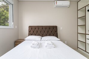 Una cama bien hecha con dos almohadas y dos toallas dobladas, una ventana con vistas a árboles, y un aire acondicionado de pared.