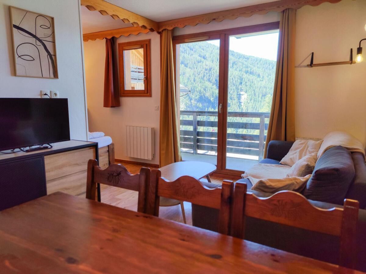 Una habitación con una mesa de comedor de madera, sillas, un sofá, un televisor y una ventana con vistas a una montaña boscosa.