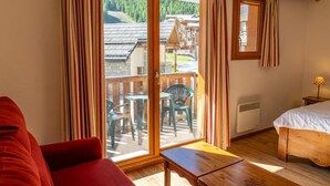 A room with a wooden bed, a red sofa, a small table, and a balcony with a view of wooden buildings and trees. - 40m from ski slopes, 1st floor, view mountain, fitness, swimming pool, sauna, balcony, tv (Les Orres)