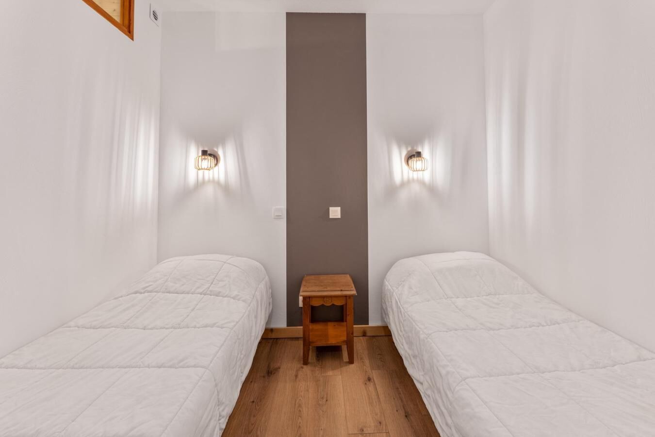 Une petite chambre propre, avec deux lits simples, une table de chevet en bois et des lampes fixées au mur.