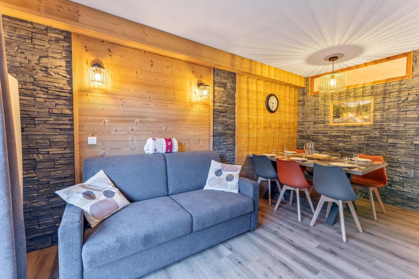 Une pièce chaleureuse avec un canapé gris, une table à manger et des chaises, des murs en pierre et une horloge au mur.