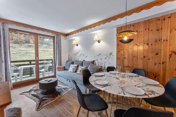 A dining area with a round table set for four, a sofa with cushions, and a view of a mountainous landscape through large windows. - Resort center, 2nd floor, view mountain, fitness, swimming pool, sauna, balcony, tv, ski locker (Les Orres)