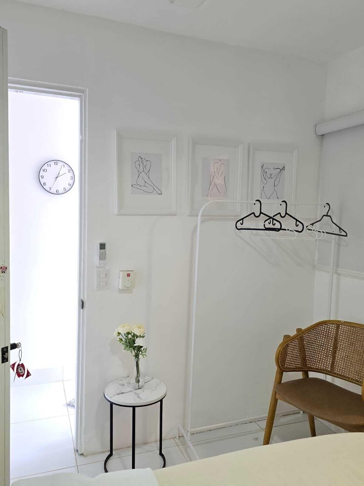 A room with a bed, a chair, a small table with a vase of flowers, a coat rack with hangers, and framed artwork on the wall.
