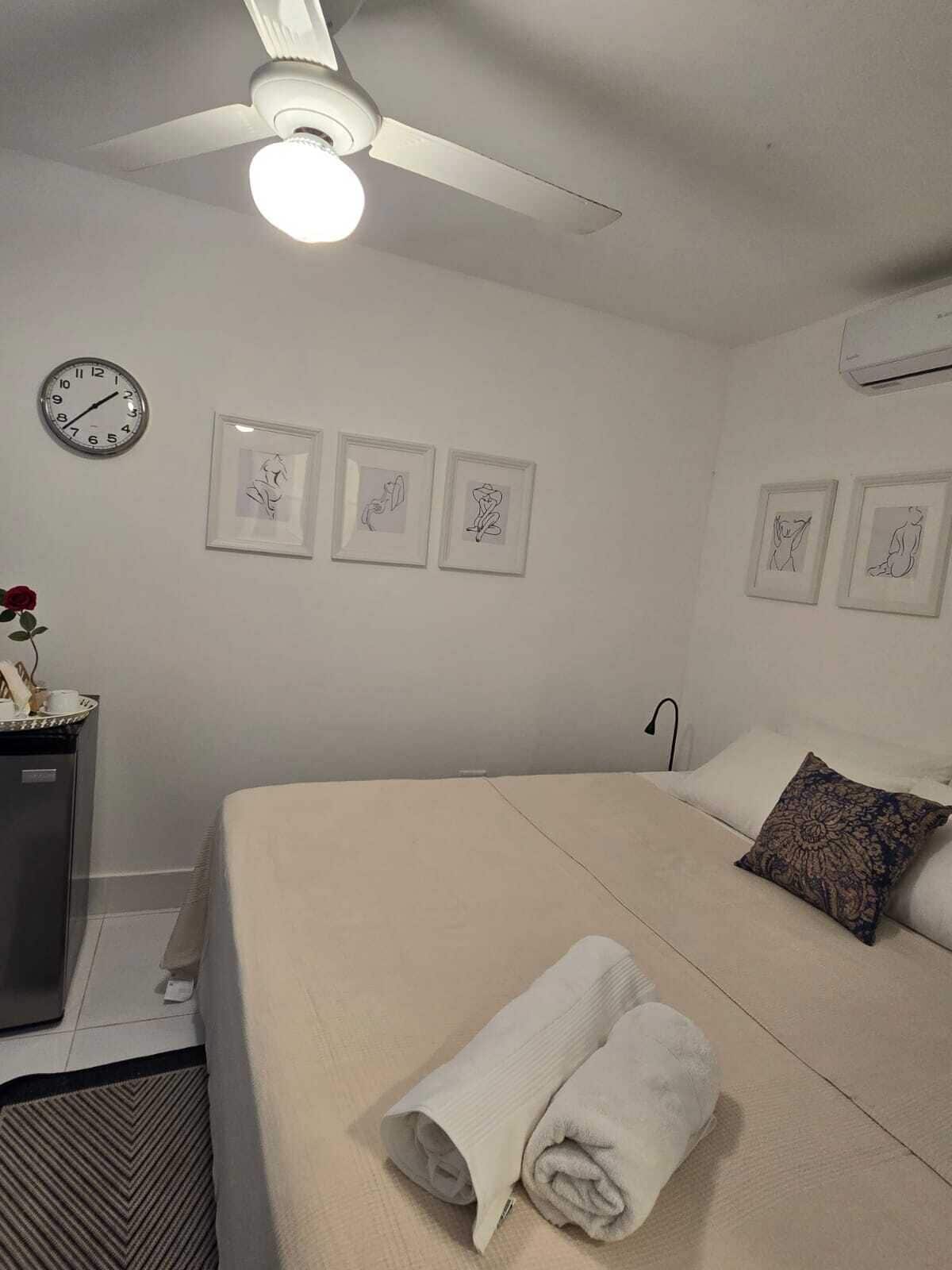 A bedroom with a bed, a ceiling fan, a clock, and framed artwork on the wall.