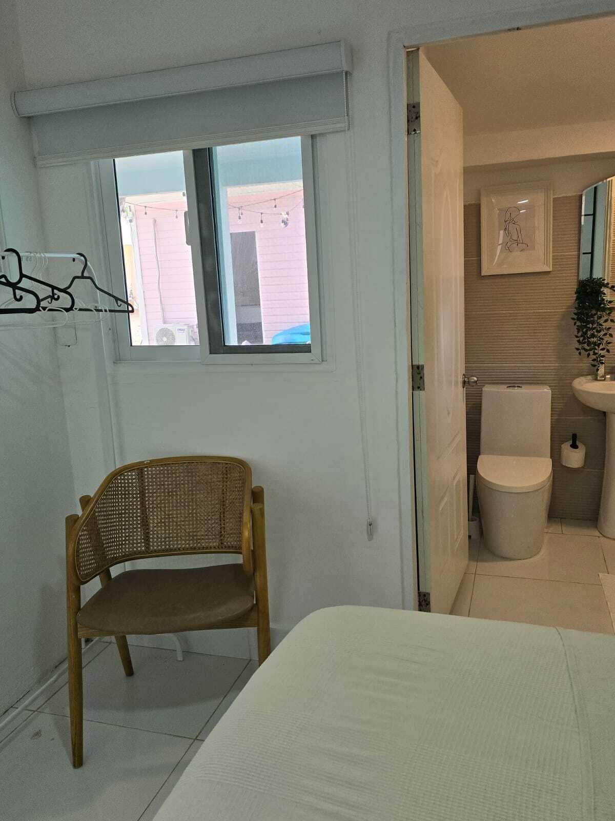Une pièce comprenant un lit, une chaise en osier, une fenêtre donnant sur un immeuble, ainsi qu’une salle de bain avec des toilettes et un lavabo.