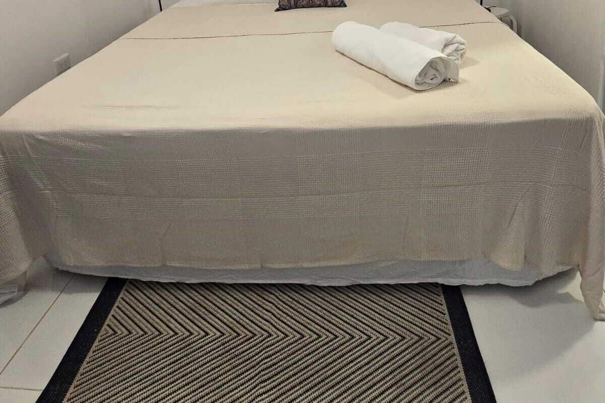 Una cama bien tendida con una colcha blanca y una toalla enrollada, una lámpara de pie negra y una alfombra estampada.