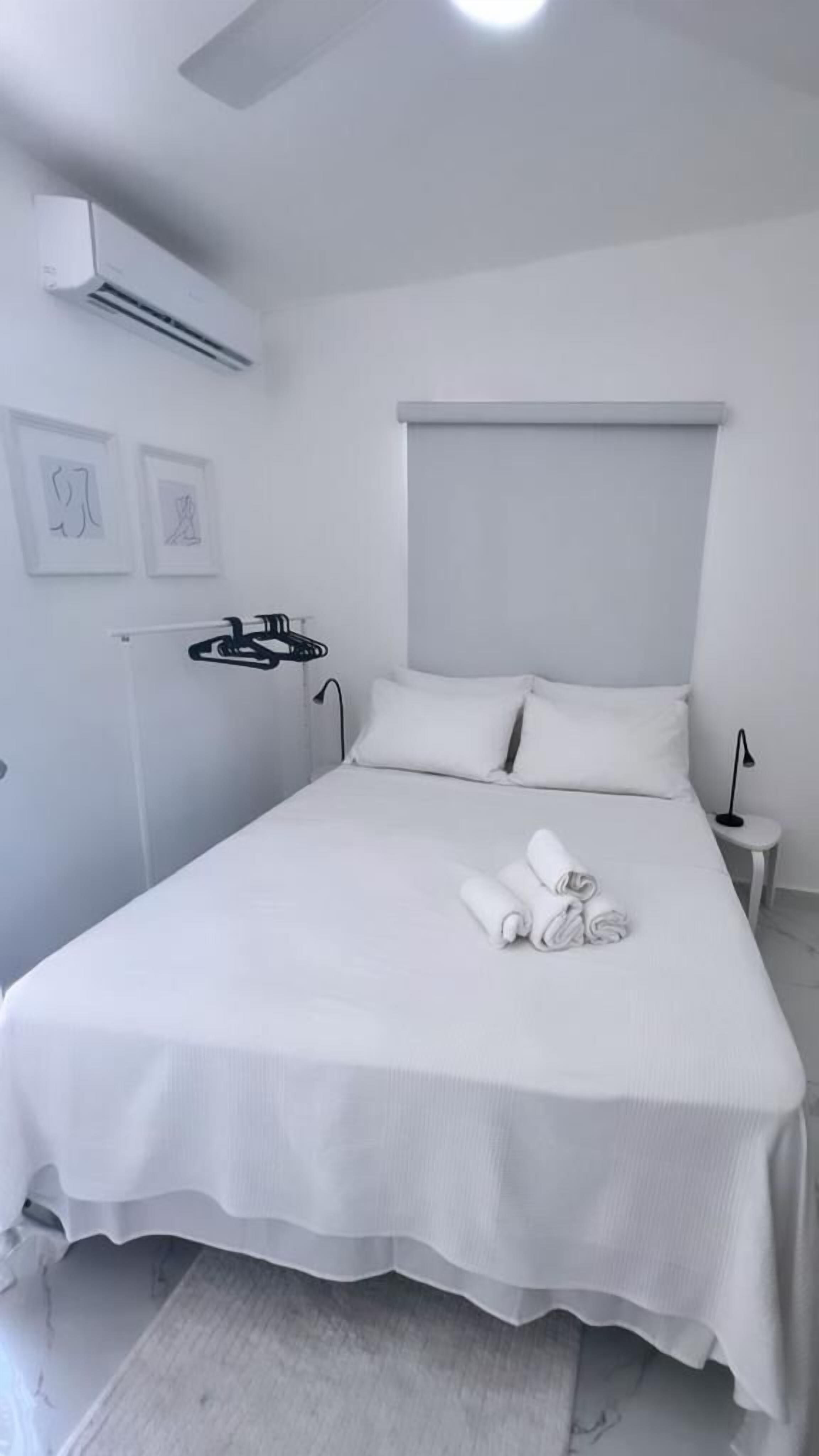 Cama bem arrumada com lençóis e toalhas brancas, luminária de cabeceira e ar-condicionado fixado na parede.