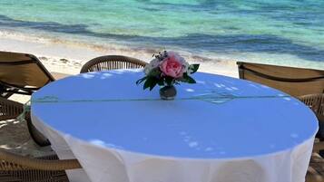 A round table with a white tablecloth and a vase of flowers, surrounded by wicker chairs, set on a sandy beach with a view of the ocean.