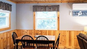 A dining area with a wooden table and chairs, a window with a checkered valance, and a wall-mounted air conditioner. - Four-Season Getaway on Upper Cold Stream Pond (Lincoln)