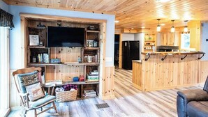 A cozy cabin interior with a wooden ceiling, a TV mounted on a wall, a kitchen area with a bar, and a seating area with a rocking chair and a sofa. - Four-Season Getaway on Upper Cold Stream Pond (Lincoln)