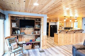 A cozy cabin interior with a wooden ceiling, a TV mounted on a wall, a kitchen area with a bar, and a seating area with a rocking chair and a sofa. - Four-Season Getaway on Upper Cold Stream Pond (Lincoln)