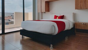 Ein modernes Schlafzimmer mit großem Fenster, einem Bett mit weißer und roter Bettwäsche, Holzschränken und Blick auf Gebäude.