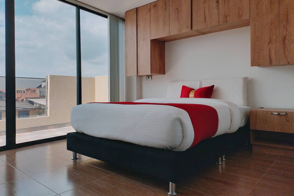 Een moderne slaapkamer met een groot raam, een bed met wit en rode beddengoed, houten kasten en een uitzicht op gebouwen.