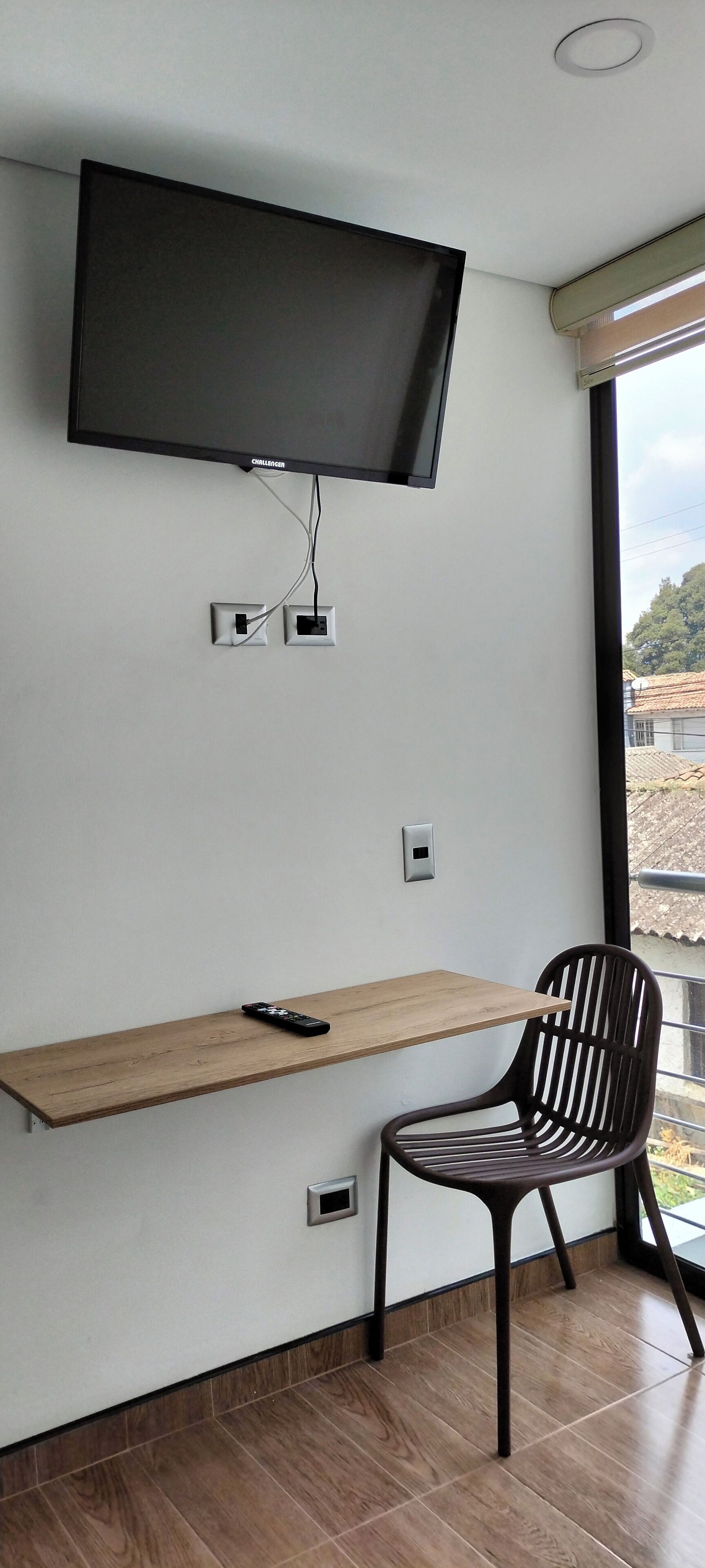 Una habitación moderna con un televisor montado, un escritorio de madera, una silla y un ventanal con vistas a edificios.
