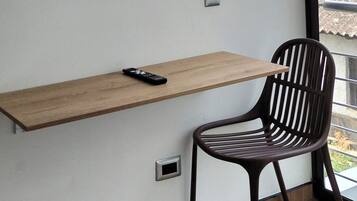 Una habitación moderna con un televisor montado, un escritorio de madera, una silla y un ventanal con vistas a edificios.