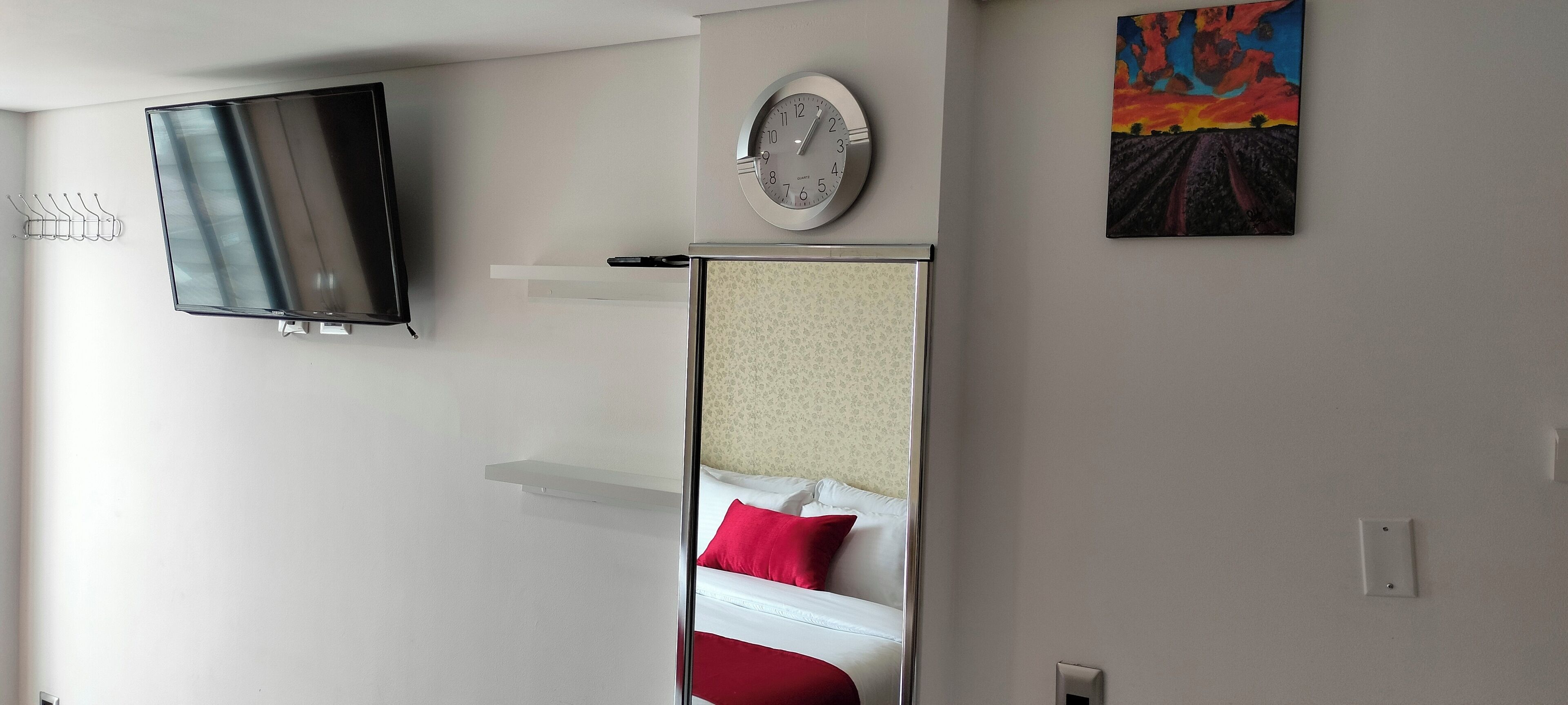 A hotel room with a flat-screen TV mounted on the wall, a clock, a painting, and a bed with red pillows.