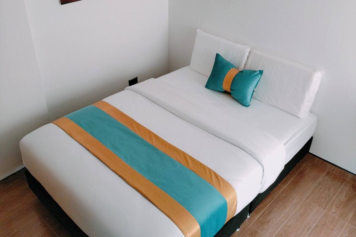 Een netjes opgemaakt bed met een witte matras, een sprei in teal- en oranjetinten en een teal-kleurig kussen.