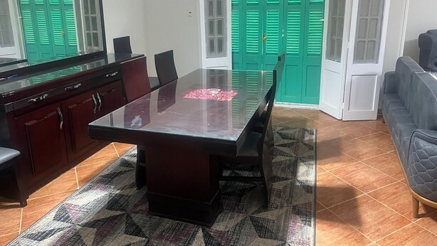 A dining area with a glass table, dark wooden chairs, a patterned rug, a sofa, and green doors in the background. - Beit Baitk 1 (6th of October City)