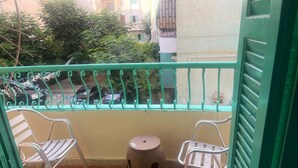 A balcony with two metal chairs and a small stool, a green railing, and a view of parked cars and greenery in the background. - Beit Baitk 1 (6th of October City)