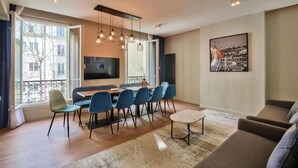 Una sala de estar moderna con comedor, ventanas amplias, televisor de pantalla plana y un cuadro de paisaje urbano.