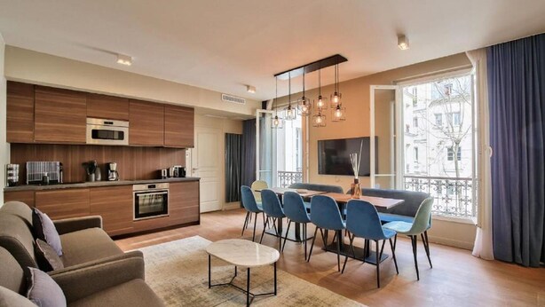 Una sala moderna con un sofá, un comedor con sillas y una mesa, una cocina con electrodomésticos, y un balcón con cortinas.