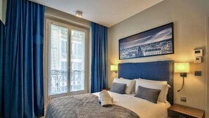 Habitación de hotel con una cama grande, un cuadro de paisaje urbano sobre la cama, un balcón con cortinas azules y una mesita de noche con una lámpara.