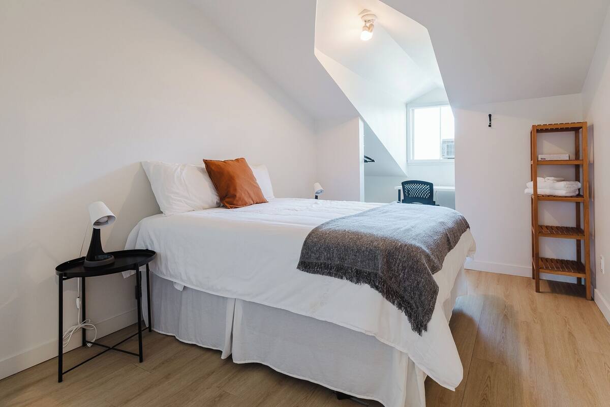 A bedroom with a bed, a bedside table, a lamp, a wooden shelf with towels, and a view of a staircase.