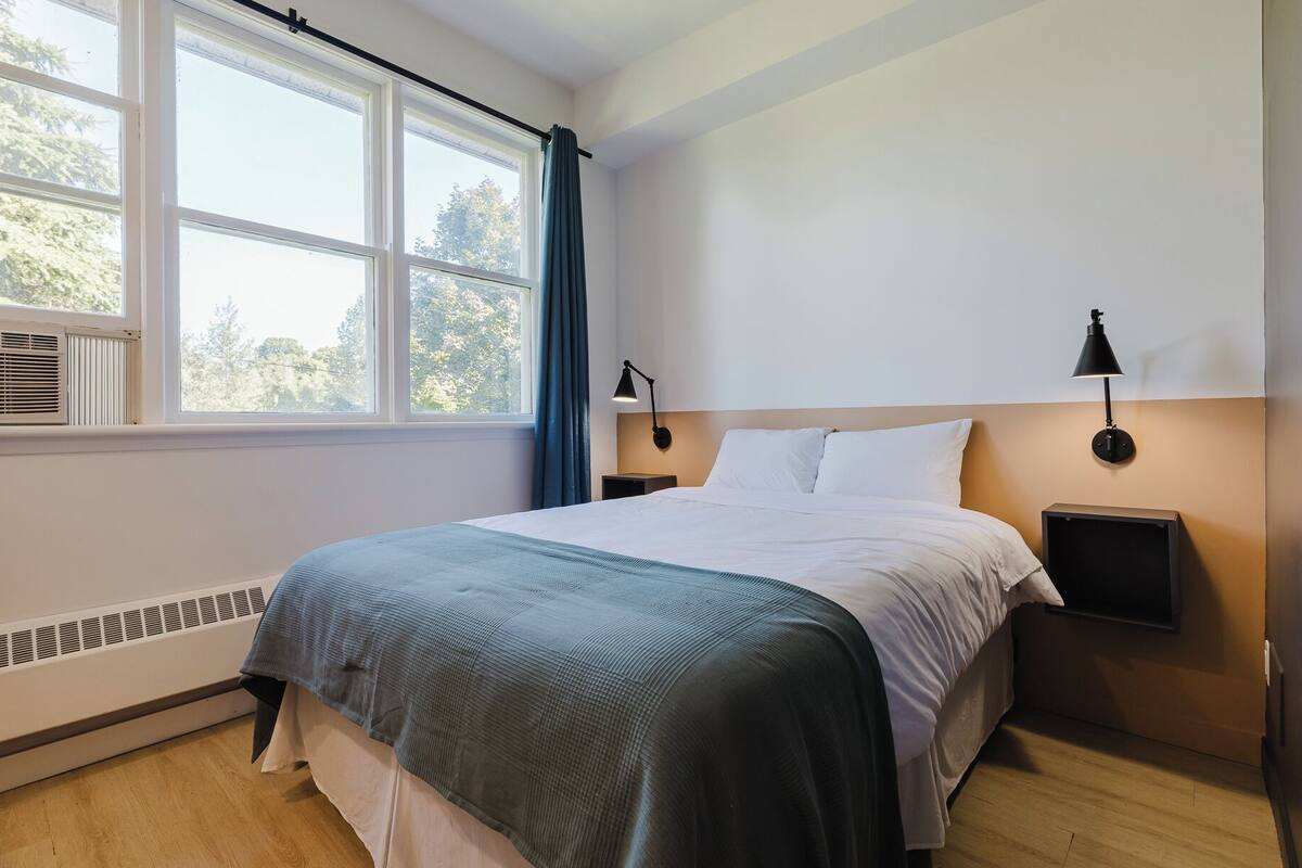 A bedroom with a bed, a window with curtains, a wall-mounted lamp, and a heater.