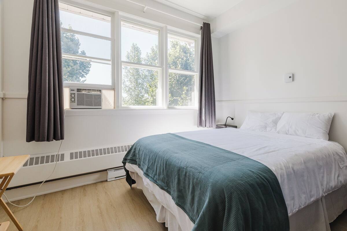 A bedroom with a bed, a window with curtains, an air conditioning unit, and a bedside table.