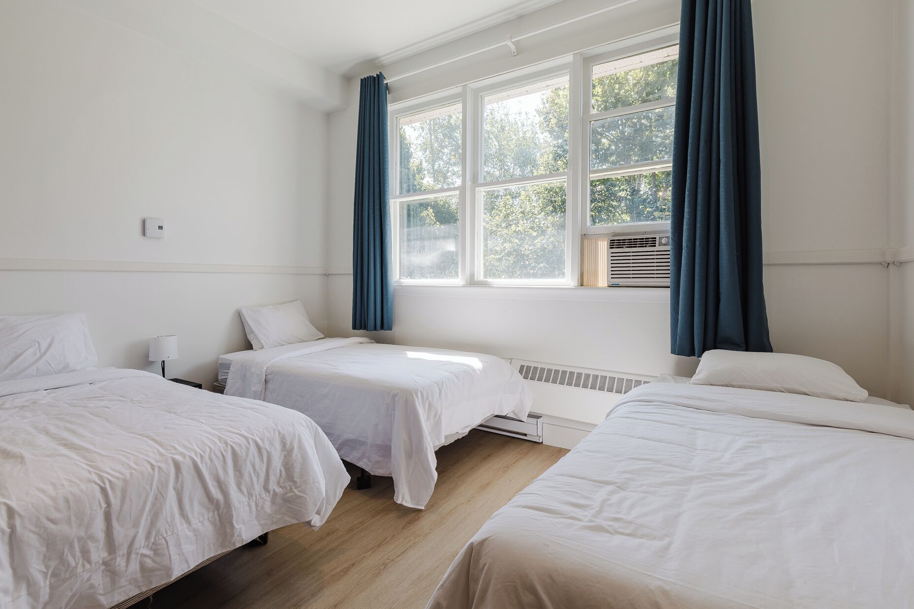A hotel room with three single beds, a window with blue curtains, and a radiator.