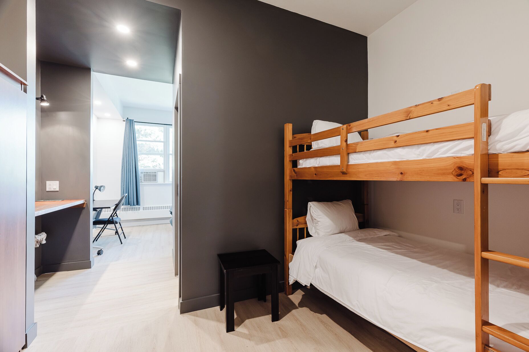 Una camera con letto a castello, una scrivania, una sedia, uno sgabello e una finestra con tende.