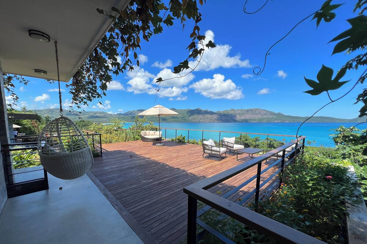 A wooden deck with outdoor furniture, a hanging chair, and a view of a clear blue sea and distant mountains.