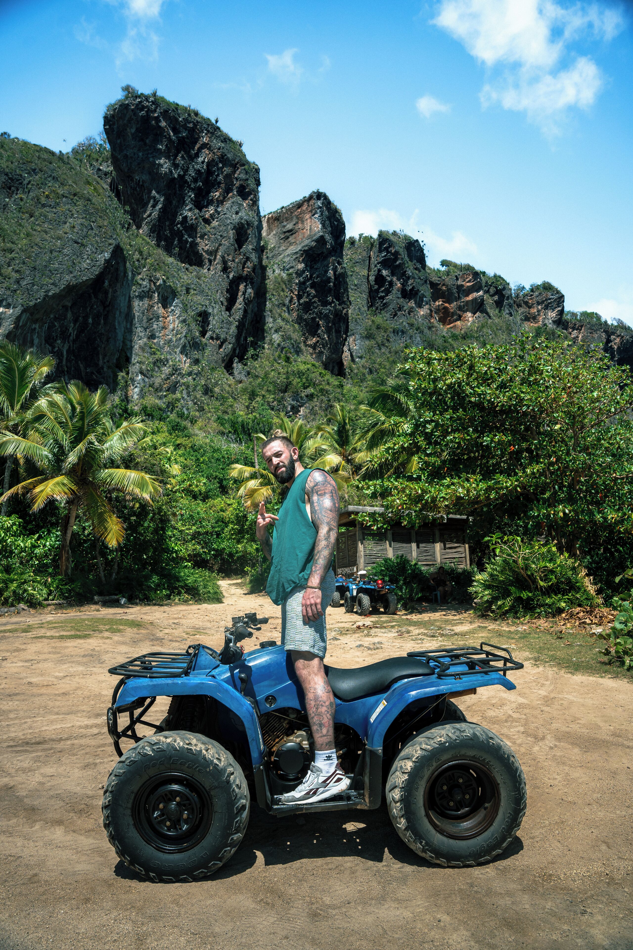 A person standing next to a blue ATV in a natural setting with palm trees and rocky cliffs in the background.