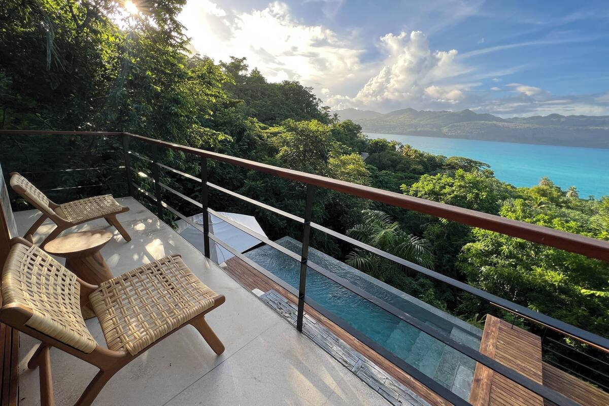 A balcony with wicker chairs and a pool overlooking a lush landscape and a clear blue sea.