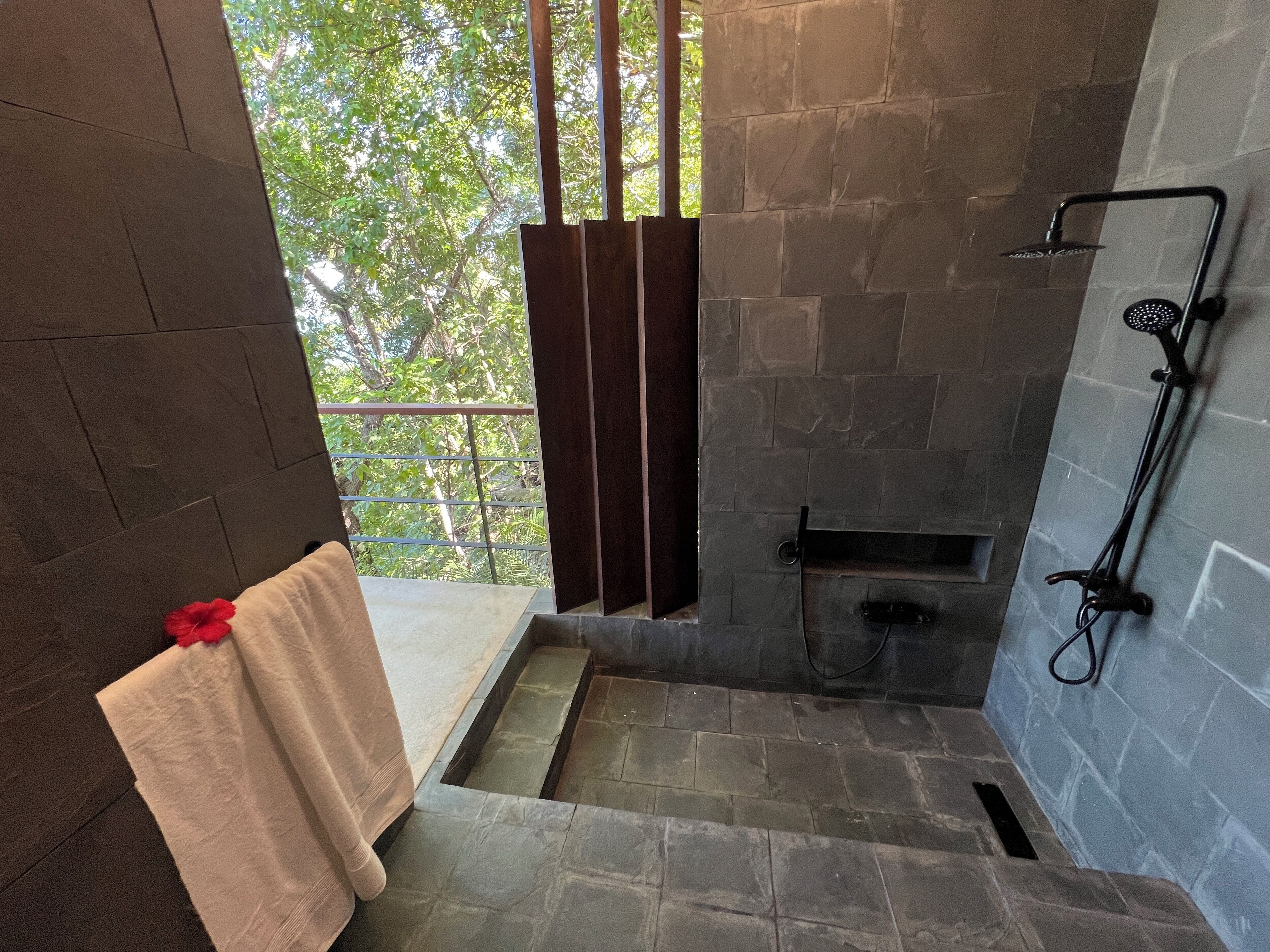 A stone-walled shower with a rain showerhead, a small bench, and a view of greenery through a window.