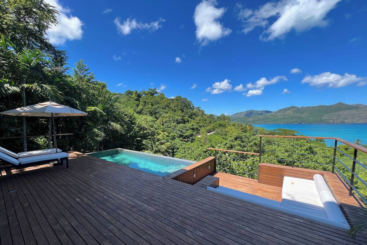 A wooden deck with a pool, lounge chairs, and a view of the ocean and mountains.
