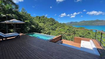 A wooden deck with a pool, lounge chairs, and a view of the ocean and mountains.