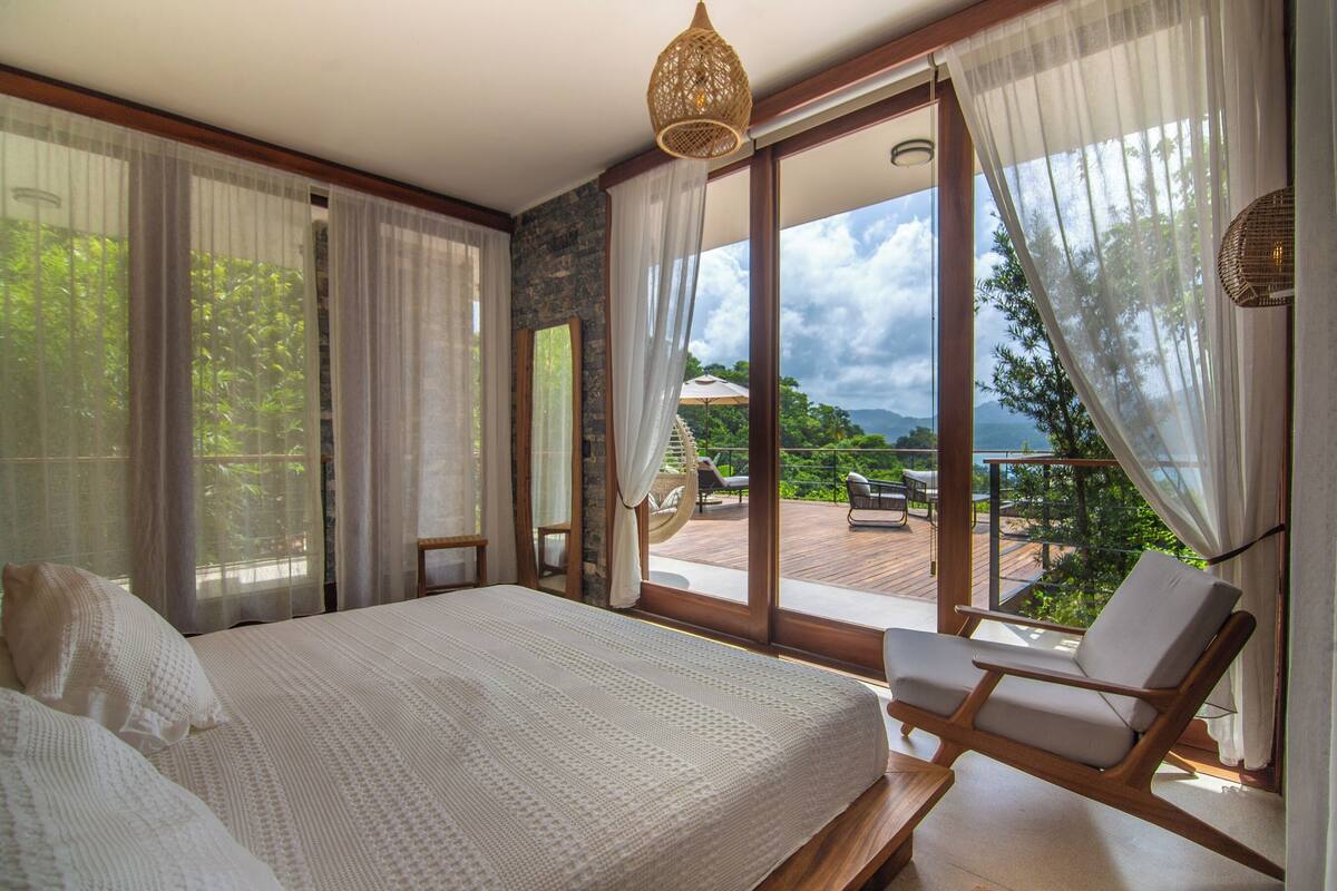 A bedroom with a large bed, a wooden chair, and a view of a deck and greenery.