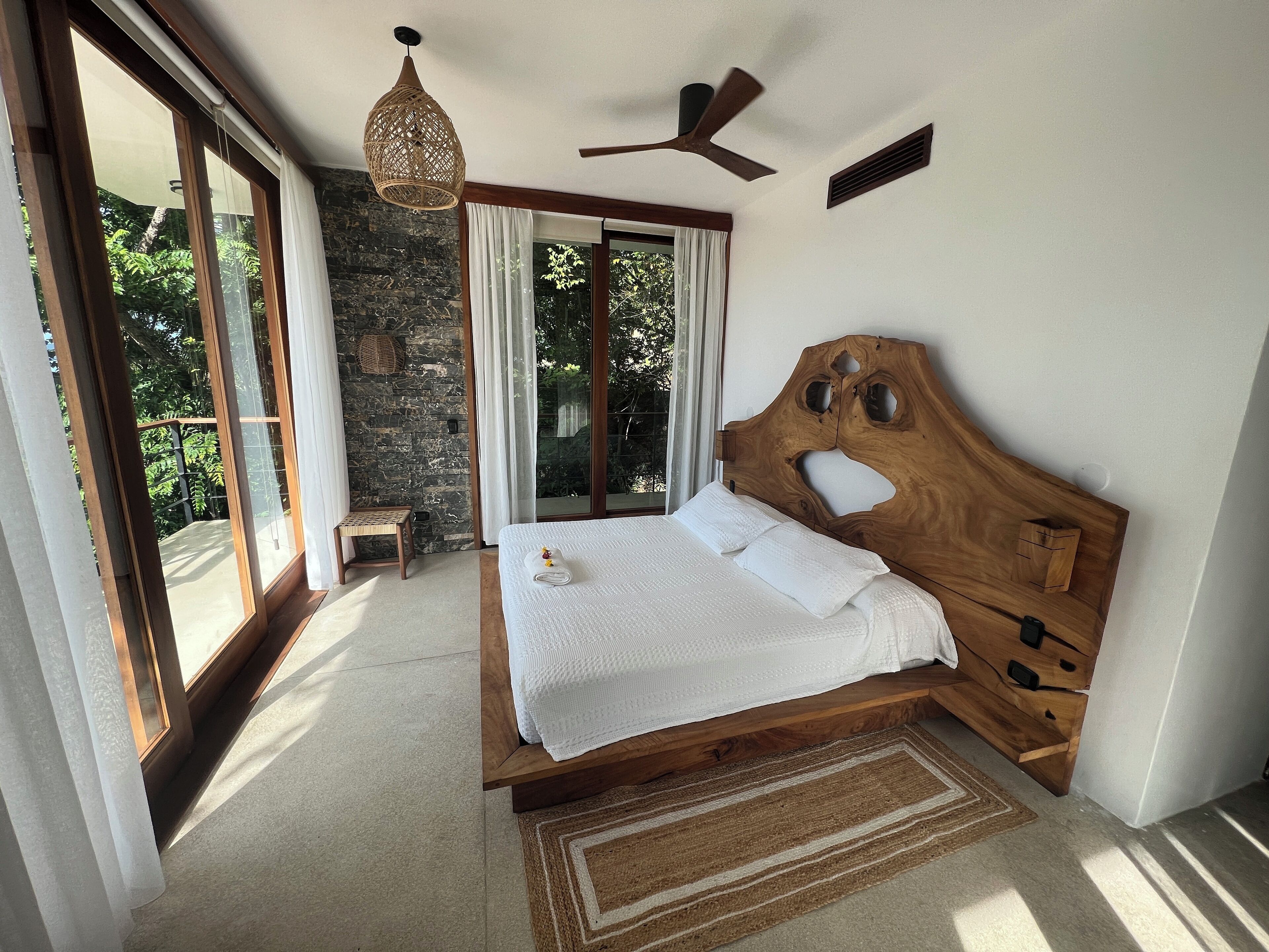 A bedroom with a wooden headboard, a bed with white linens, a small wooden stool, a woven light fixture, and a view of greenery through large windows.