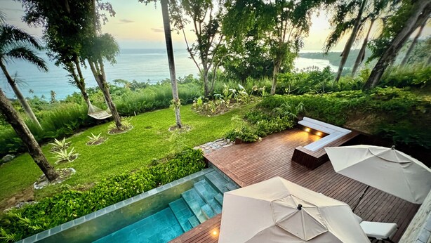 A swimming pool with a wooden deck, surrounded by lush greenery and palm trees, overlooking a body of water. - Ocama (Las Galeras)