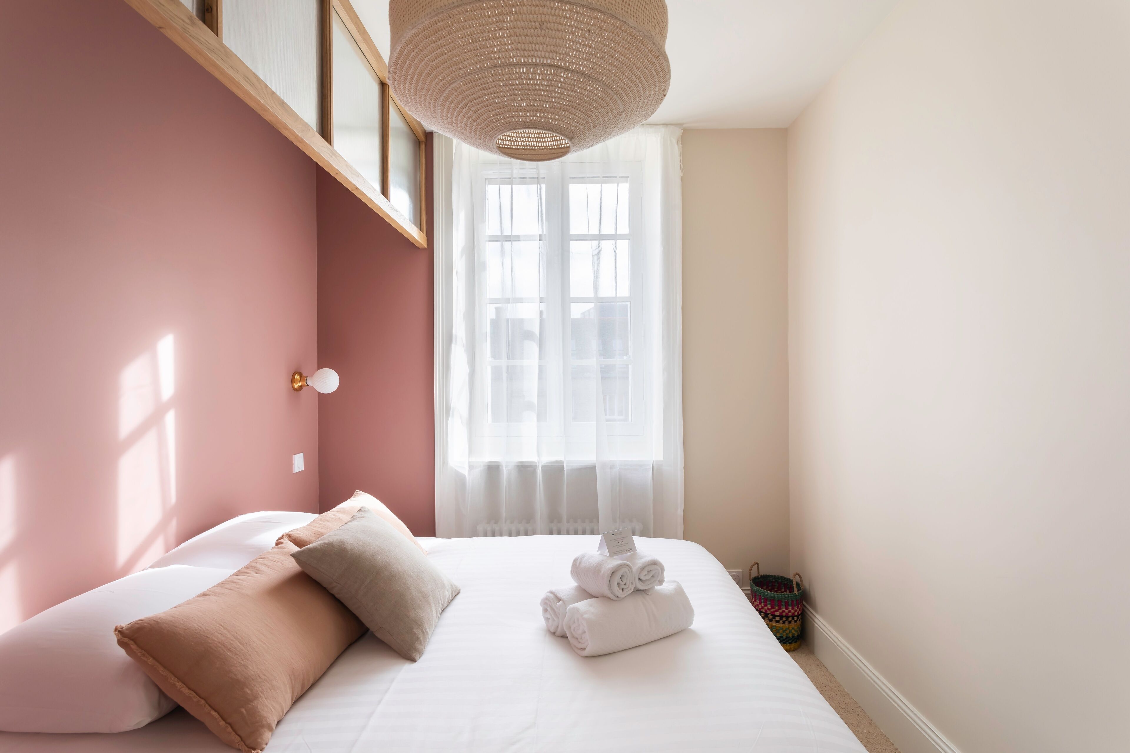 Un dormitorio con cama, almohadas, una ventana con cortinas, una lámpara colgante y una cesta pequeña.