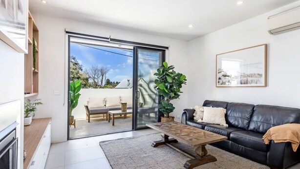 Una sala de estar moderna con un ventanal corredero que da a un patio exterior, un sofá de cuero negro, una mesa de centro de madera y una planta grande en maceta.