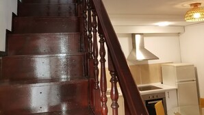 A wooden staircase with a handrail, leading to a room with a kitchen area, a dining table with chairs, and a lamp on a dresser. - Casa Cadu - Porto (Porto)